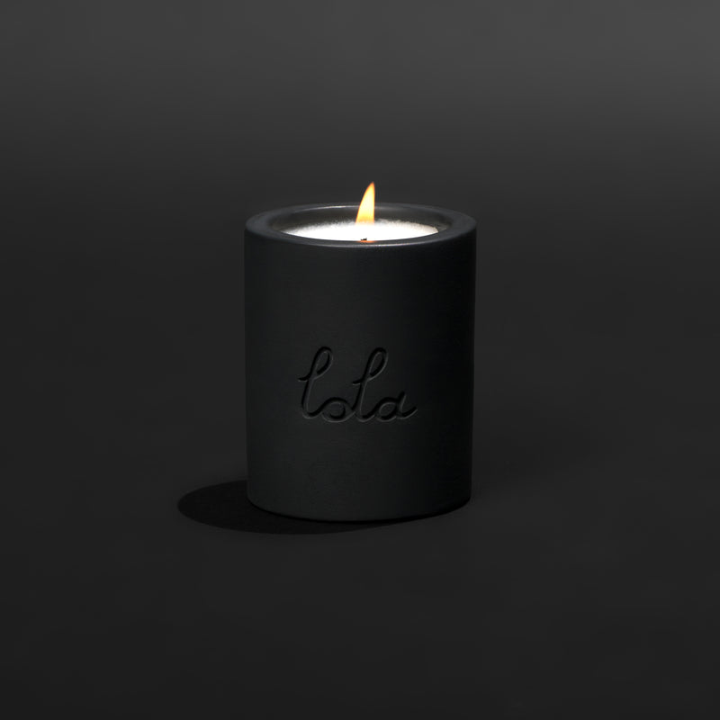 Three cylindrical candles with lit flames, placed side by side. From left to right, the candles are beige, white, and black, each with the brand name 'Lola' written on the front.