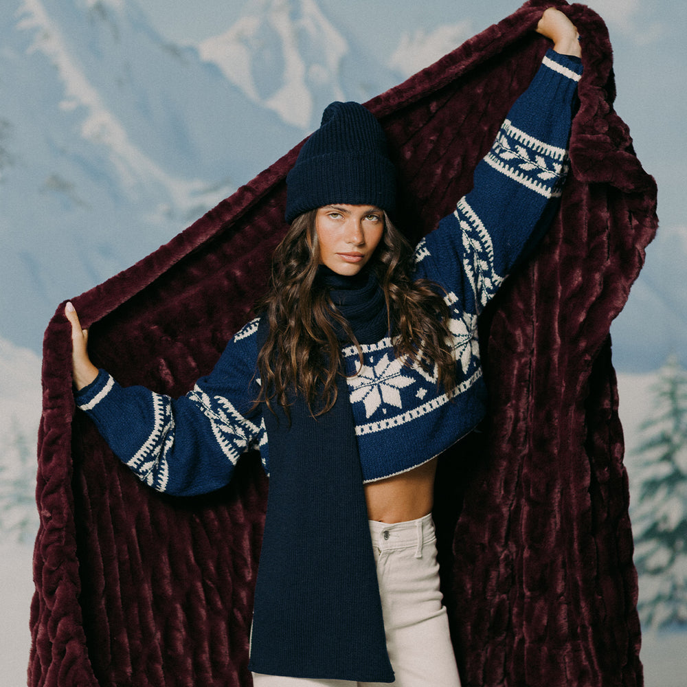 Woman wearing a blue sweater with white patterns, holding a dark red blanket against a mountainous background.