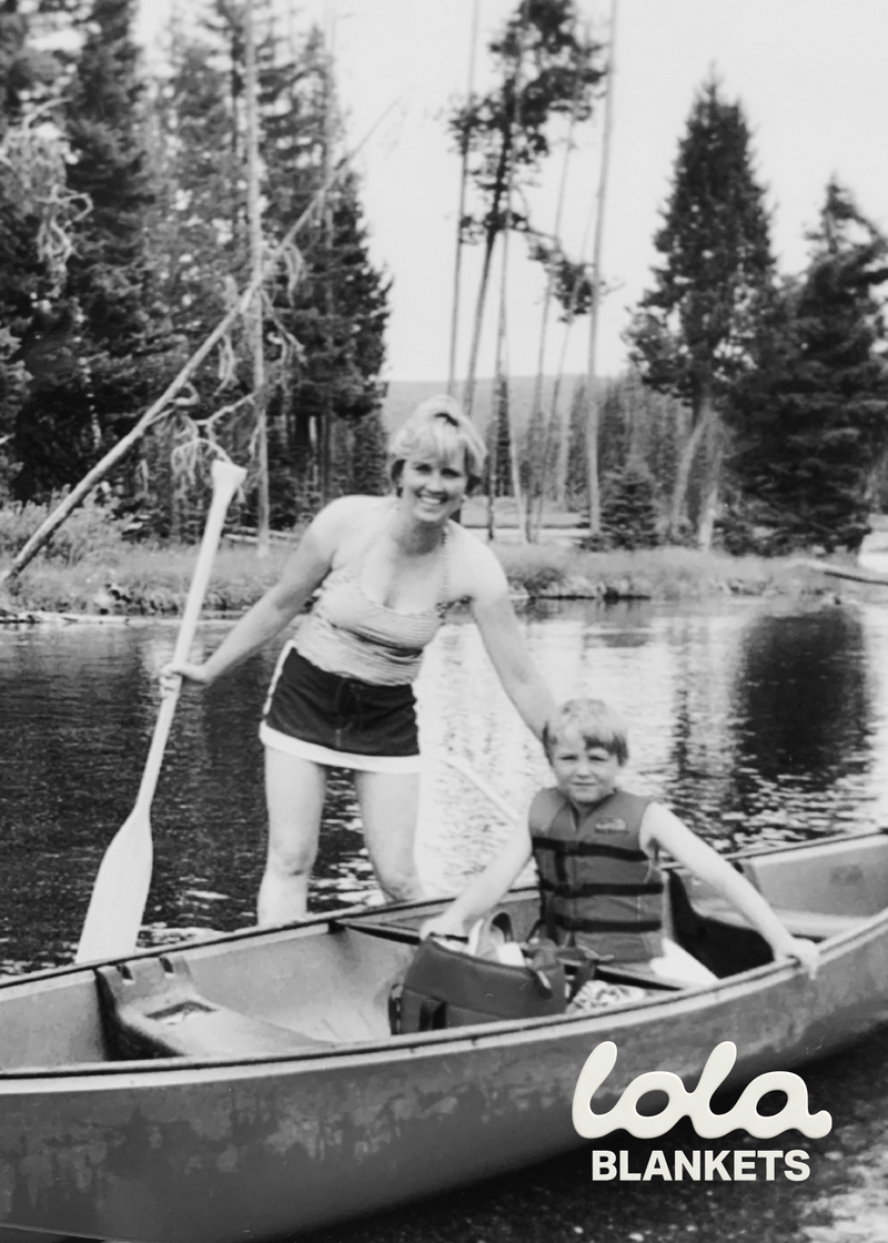 Black and white photo of a woman and child in a canoe with trees and water in the background, featuring 'lola BLANKETS' branding.