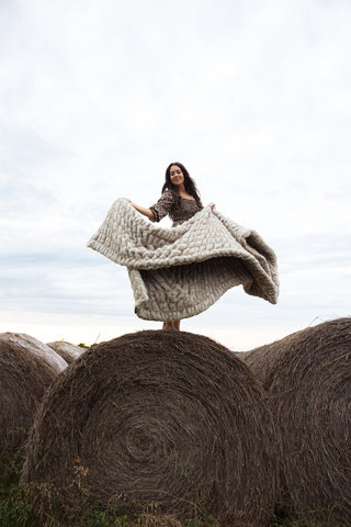 Woman holding a large textured blanket over her shoulders in an outdoor setting with hay bales.