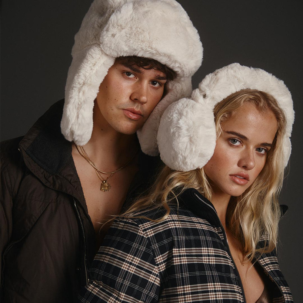 Two people wearing white fluffy bomber hats and fluffy ear muffs against a dark background