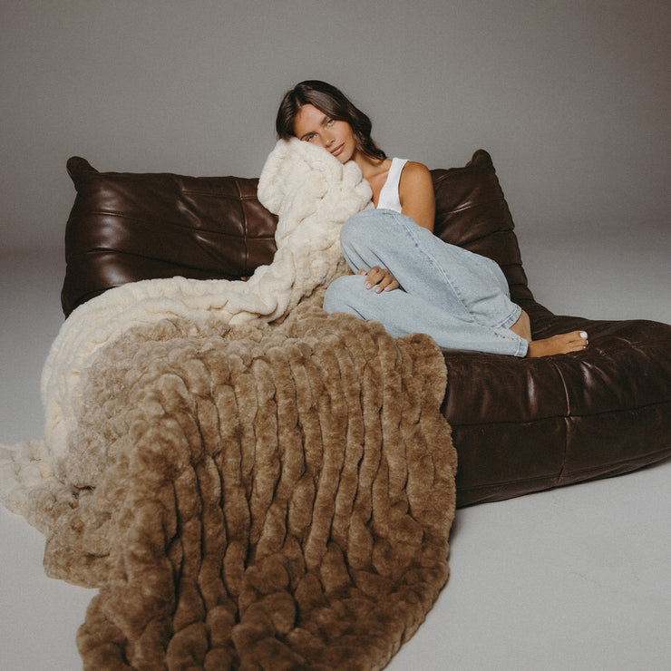 Woman sitting on a brown leather couch with a textured blanket draped over her.