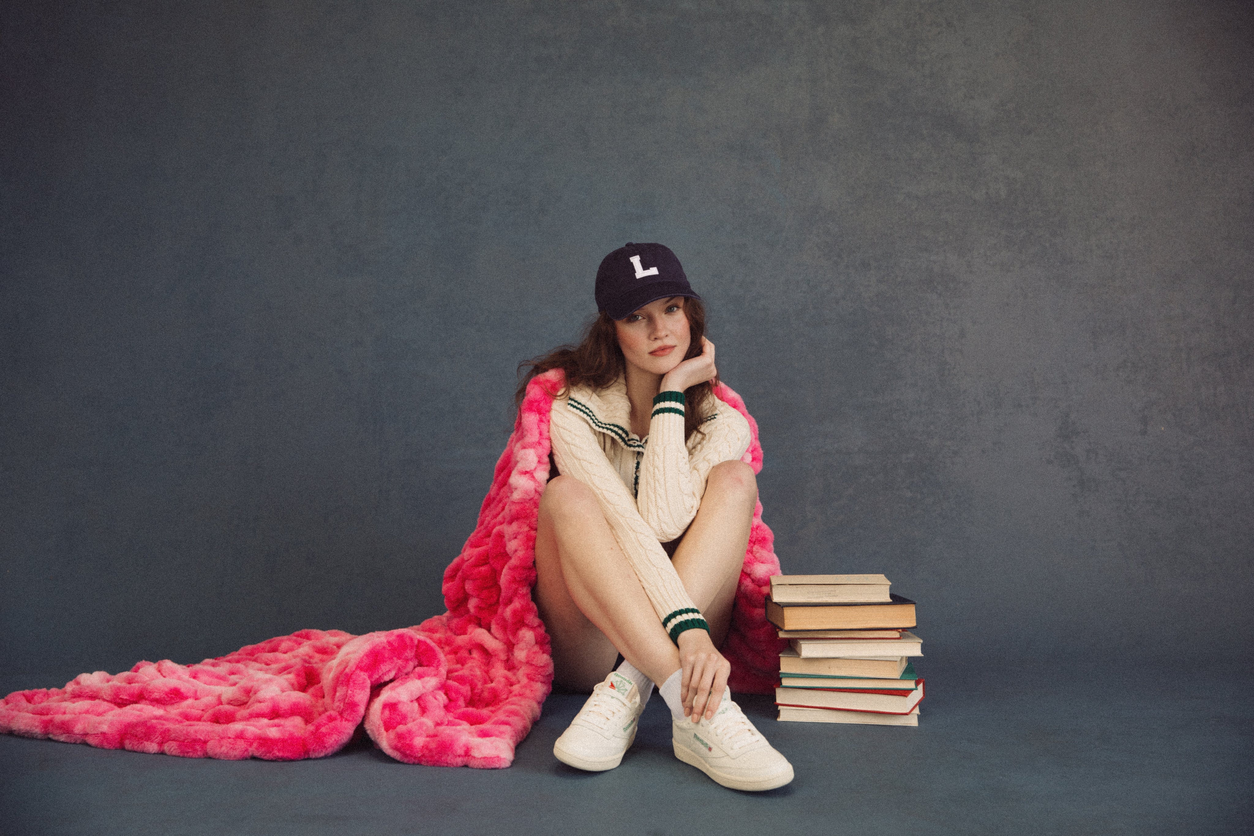 Person sitting on a stack of books with a pink blanket and wearing a black cap against a dark background