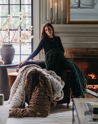 A woman sitting on the arm of a sofa, showcasing textured Lola blankets.