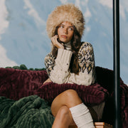 Woman wearing a fur hat and patterned sweater sitting on a couch with a scenic background