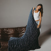 Woman holding a large blue textured blanket in front of a brown leather couch.