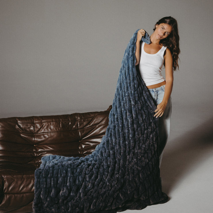 Woman holding a large blue textured blanket in front of a brown leather couch.