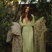 Woman in a light green dress standing between two beige knitted cardigans with a natural outdoor background.