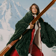 Person in a green fur coat holding skis with a mountainous background